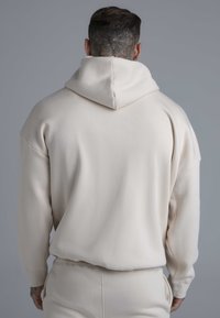 Beiger übergroßer Hoodie mit Kapuze, der eine weiche Textur und lange Ärmel aufweist. Die Rückseite zeigt ein minimalistisches Design ohne sichtbare Logos.