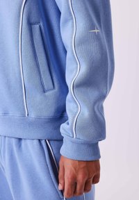 Lichtblauwe sweatshirt met een geribde zoom en manchetten, voorzien van witte bies langs de mouwen en een kleine sterafbeelding op de arm.