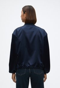 Jūras flotes satīna bomber jakas ar brīvu piegriezumu, elastīgu apakšmalu un aprocēm, ar augstu apkakli un gludu tekstūru. Attēlots aizmugures skats.