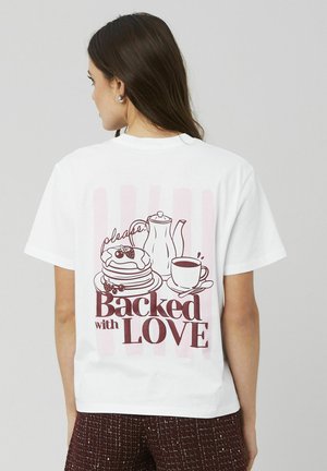 T-shirt en coton blanc avec un motif imprimé représentant des pancakes, une théière et une tasse, agrémenté de texte rose et bourgogne qui dit "Soutenu par l'AMOUR."