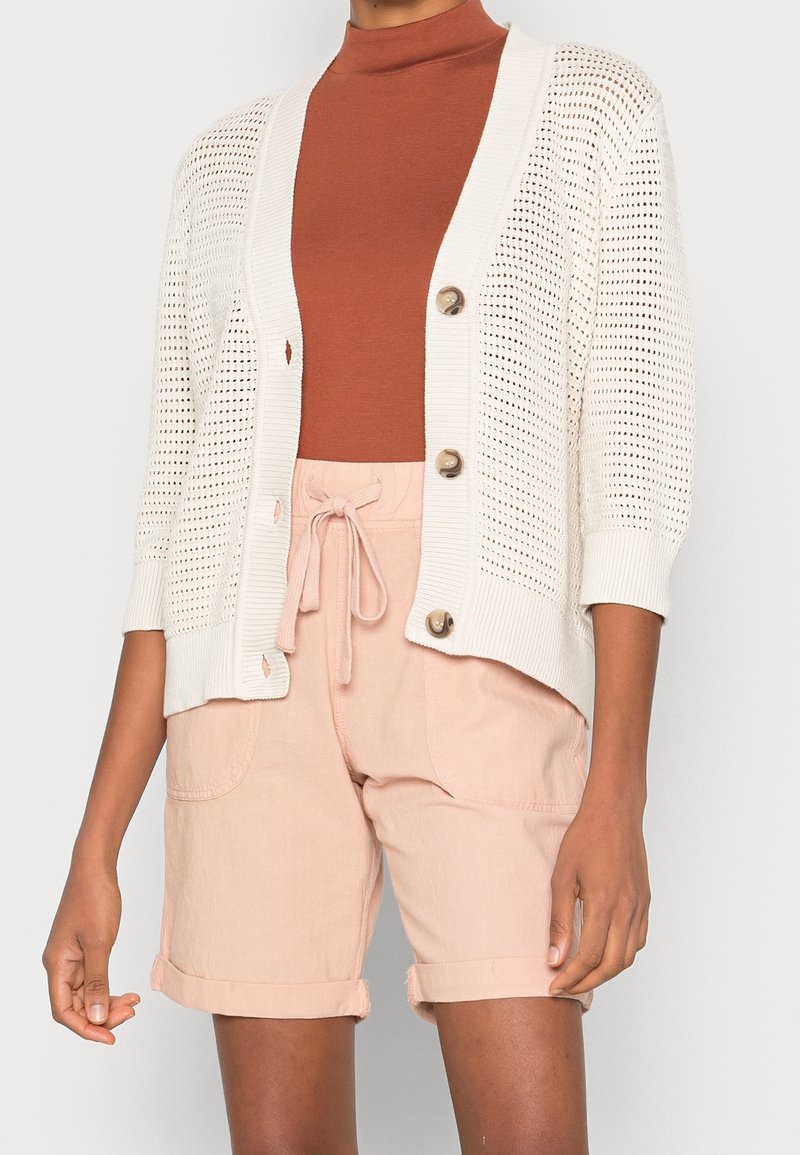 Femme portant un cardigan beige en maille ouverte avec des boutons par-dessus un col roulé rouille et un short rose clair à cordons.