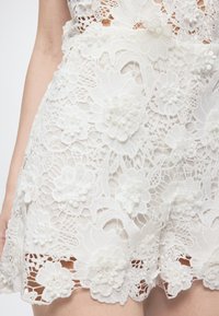 Λευκό δαντελένιο ένδυμα με περίπλοκα λουλουδένια σχέδια και υφασμένα τρισδιάστατα λουλούδια, που απεικονίζει μέρος του κορμού και του βραχίονα ενός ατόμου.