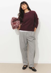 Pull en tricot bourgogne, pantalon gray à rayures fines, ballerines noires à motif, tenant un sac paisley rouge et blanc. Fond uni.