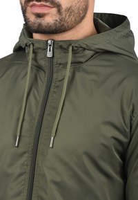 Veste à capuche vert olive avec une texture lisse, dotée d'une fermeture éclair centrale, de cordons de serrage ajustables et d'accents métalliques près de la fermeture éclair.