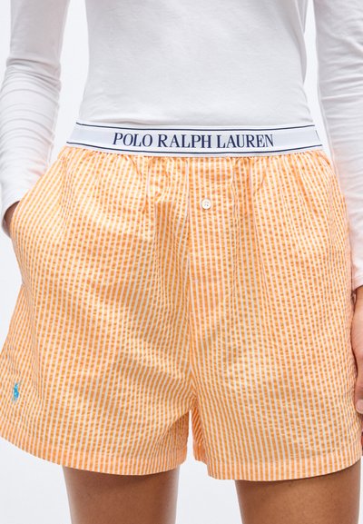 Άτομο που φορά πορτοκαλί και λευκά ριγέ σορτς με ζωνάρι με την επωνυμία Polo Ralph Lauren και έχει τα χέρια στις τσέπες.