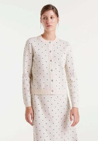 Cárdigan de crema claro con lunares negros, con botones en el frente, puños y dobladillo acanalados. Combínalo con una falda de patrón similar para un look coordinado.