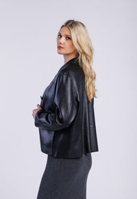 Giacca in pelle nera con vestibilità rilassata e apertura frontale. Presenta un colletto, maniche lunghe e una texture liscia. Indossata sopra una gonna grigia.
