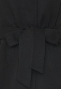 Schwarzer Strickstoff mit einem gebundenen Gürtel-Detail in der Taille. Die Textur wirkt weich und das Design weist eine glatte, gleichmäßige Oberfläche auf.