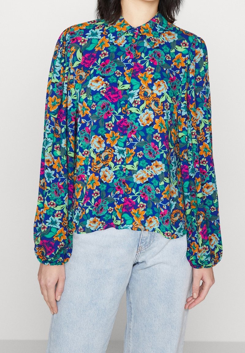 Blouse à manches longues à motifs floraux sur fond bleu, avec des fleurs vives orange, rose et vertes. Col boutonné et coupe ample.