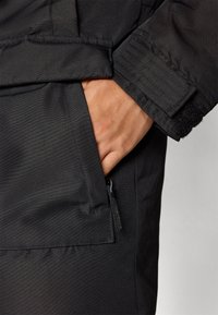 Chaqueta negra de tejido suave. Presenta un bolsillo con cremallera y una mano en el bolsillo lateral. La manga tiene un acento de canalé.