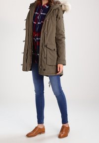 Olivgrön parkerjacka med en huva av syntetisk päls, framsida med knappar och fickor, kombinerad med en rutig skjorta, tajta jeans och bruna snörskor.