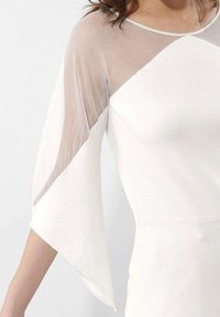 Witte jurk met doorschijnende mesh mouwen en een aansluitende lijfje. De mouwen zijn aan de uiteinden wijd uitlopend, wat contrast toevoegt aan de stevige stof.