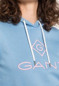 Lichtblauwe hoodie met een trekkoord, voorzien van een roze geborduurd logo en merknaam "GANT" op de voorkant. Zachte stoftextuur.