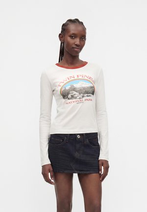 Kvinde med flettet hår iført en hvid langærmet skjorte med Twin Pines National Park-print og en mørk denim mininederdel, står foran en ensfarvet baggrund.