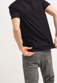 Persona con camiseta negra y jeans grises rascándose la parte baja de la espalda con la mano derecha, levantando ligeramente la camiseta con la mano izquierda, contra un fondo plano.