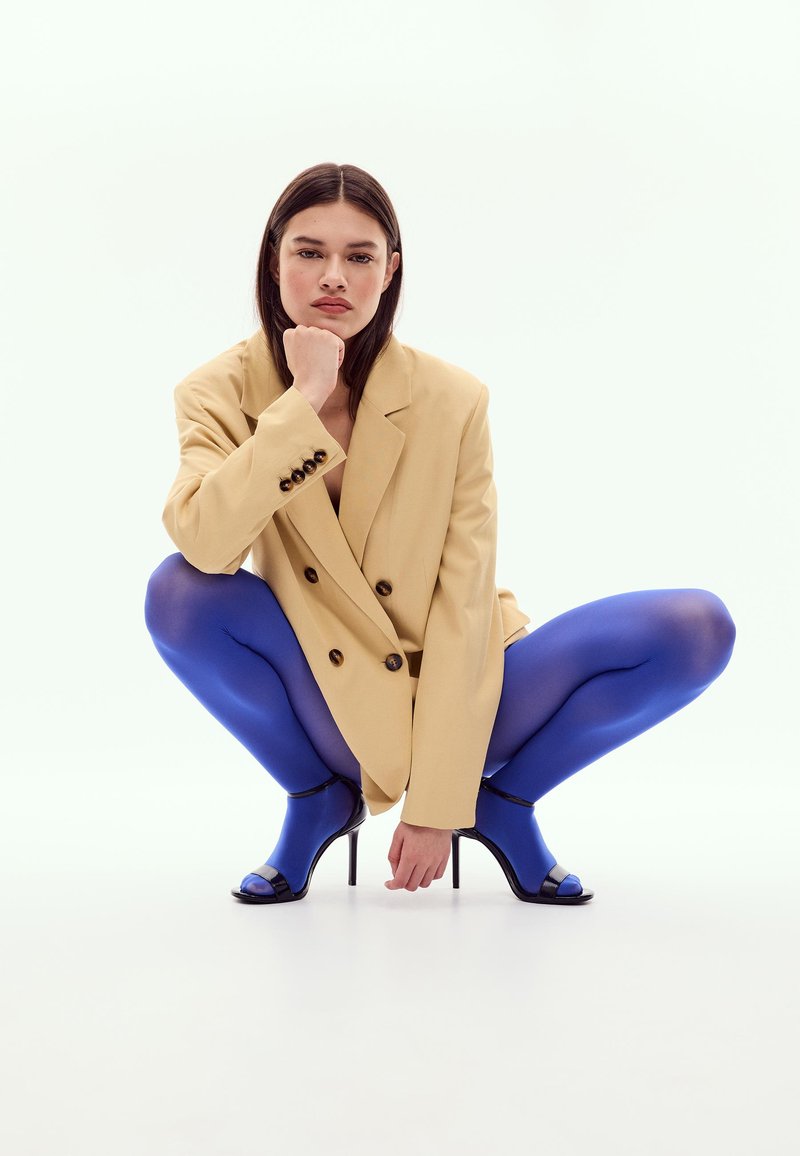 Femme accroupie portant un blazer oversize beige, des collants bleu vif et des chaussures à talons hauts noires ouvertes à l'avant sur fond blanc.