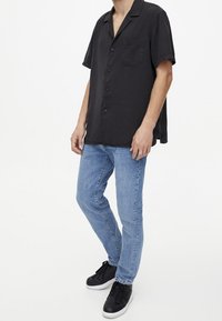 Chemise noire à manches courtes avec boutons et poche, associée à un jean bleu clair et des baskets noires. Tissu lisse, coupe décontractée.
