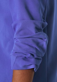 Detailní pohled na fialovou košilovou manžetu, která zobrazuje měkkou látku s ohrnutým rukávem, zdůrazňující texturu a barvu.