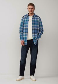 Koszula w wielobarwną kratę w niebieskich i zielonych odcieniach, noszona na białej bluzie henley, w zestawieniu z ciemnymi dżinsami i białymi trampkami.