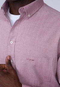 Chemise rose pâle à boutons en tissu tissé. Elle présente un col classique, une poche frontale avec détail de couture, et une patte de boutonnage.