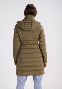 Žena oblečená v olivově zeleném prošívaném kabátu s kapucí a páskem, stojící zády, doplněná o světle modré džíny.