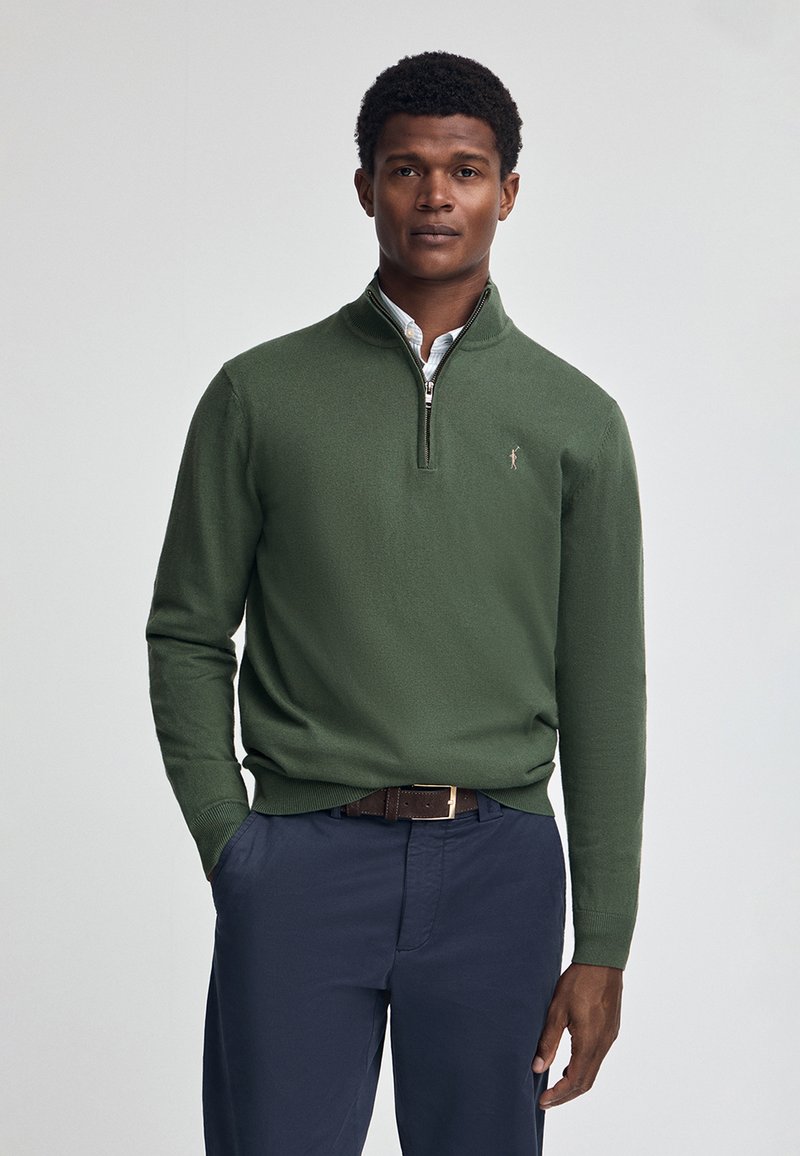 Mörkgrön pullovertröja med dragkedja i kragen, tillverkad av mjukt material, med en liten logotyp på vänster bröst och ribbad nedkant och ärmslut.