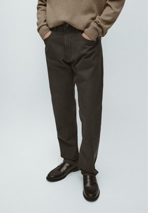 Person trägt dunkelbraune Hose, braunen Pullover mit den Händen in den Taschen und schwarze Lederschuhe, steht vor einem schlichten hellen Hintergrund.