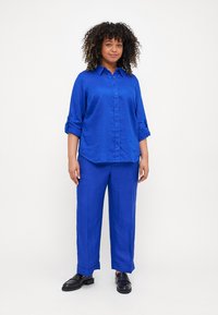 Camisa azul clara de botão de manga longa com mangas arregaçadas, combinada com calças largas azuis e sapatos de loafers pretos. Textura suave.