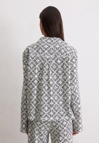 Mujer con cabello largo y oscuro, vistiendo una camisa blanca y pantalones con un patrón cuadrado floral negro, de pie contra una pared lisa clara, de espaldas a la cámara.