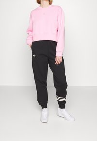 Osoba v světle růžové mikině a černých kalhotách Adidas se bílými pruhy, stojící s rukama v kapsách, v bílých teniskách.