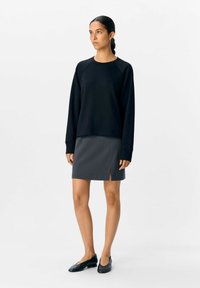 Sorte sweatshirt med raglanærmer, parret med en grå mini-nederdel med en sideslids, båret med sorte flade sko på en enkel baggrund.