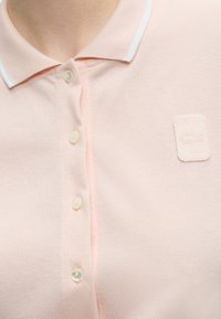 Polo rose clair avec col bordé de blanc, quatre boutons et un petit écusson brodé avec logo sur la poitrine.