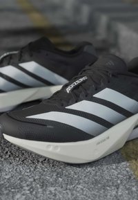 Chaussures de course Adidas Adizero noires et blanches avec semelles Lightstrike Pro sur une surface grise texturée.