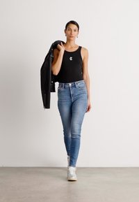 Czarny top na ramiączkach z logo, jasnoniebieskie wąskie dżinsy i białe sneakersy. Model trzyma czarną skórzaną kurtkę. Jasne tło i gładka podłoga.