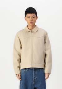Chaqueta beige con cremallera y cuello de pana, de mangas largas y corte relajado, combinada con pantalones de mezclilla azul holgados. Textura suave, diseño minimalista.