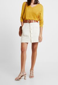 Vrouw in een mosterdgele blouse met lange mouwen, witte rok met knopen aan de voorkant, bruine riem en tas, en beige sandaaltjes met blokhak, staand op een witte vloer.