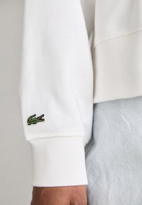 Hanorac alb cu manșete ribbate, având un logo brodat în formă de crocodil verde pe mâneca stângă. Textură moale și croi lejer.