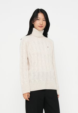 Woman with long black hair wearing a cream cable-knit turtleneck sweater and black pants against a white background.