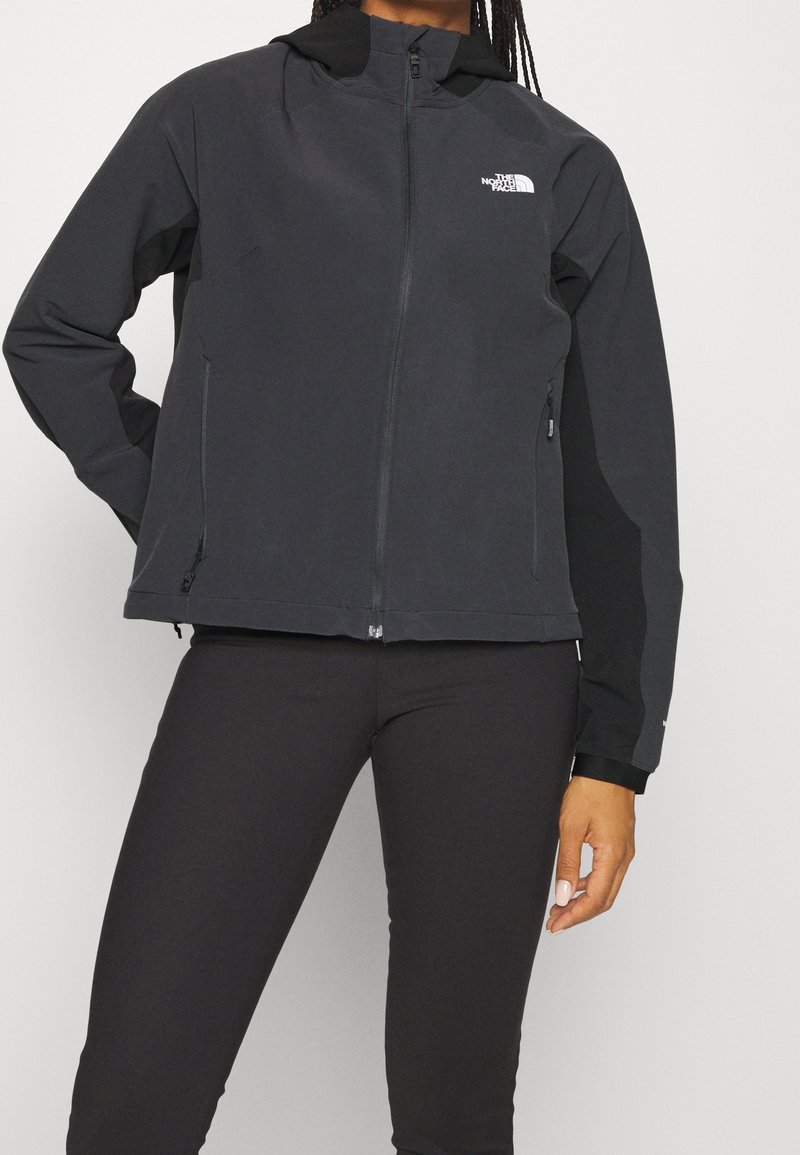 Sort softshell-jakke med aftagelig hætte, lynlåslukning foran, sidelommer og et logo på venstre bryst, med en glat tekstur.