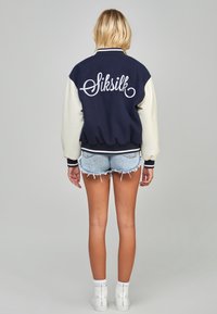 Marinblå och krämfärgad varsityjacka med ribbad mudd, med en vit broderad logotyp på baksidan, tillsammans med slitna denimshorts och vita sneakers.