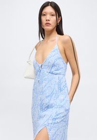 Νεαρή γυναίκα με ίσια μαύρα μαλλιά που φοράει ανοιχτά μπλε φόρεμα με σχέδιο παϊέτες και κρατάει άσπρη τσάντα στον ώμο.