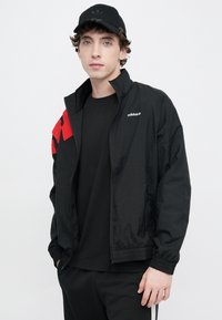 Μαύρο ελαφρύ μπουφάν με κόκκινο και μαύρο μοτίβο στον ώμο, με φερμουάρ κλεισίματος και ελαστικές μανσέτες. Λογότυπο τυπωμένο στο στήθος.