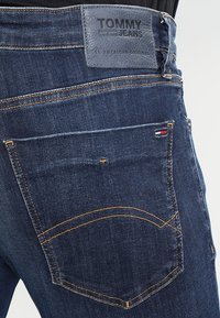 Mörkblå denimjeans med en läderlogotyp som är märkt "TOMMY JEANS", kontrasterande sömmar och en distinkt design på bakfickan.