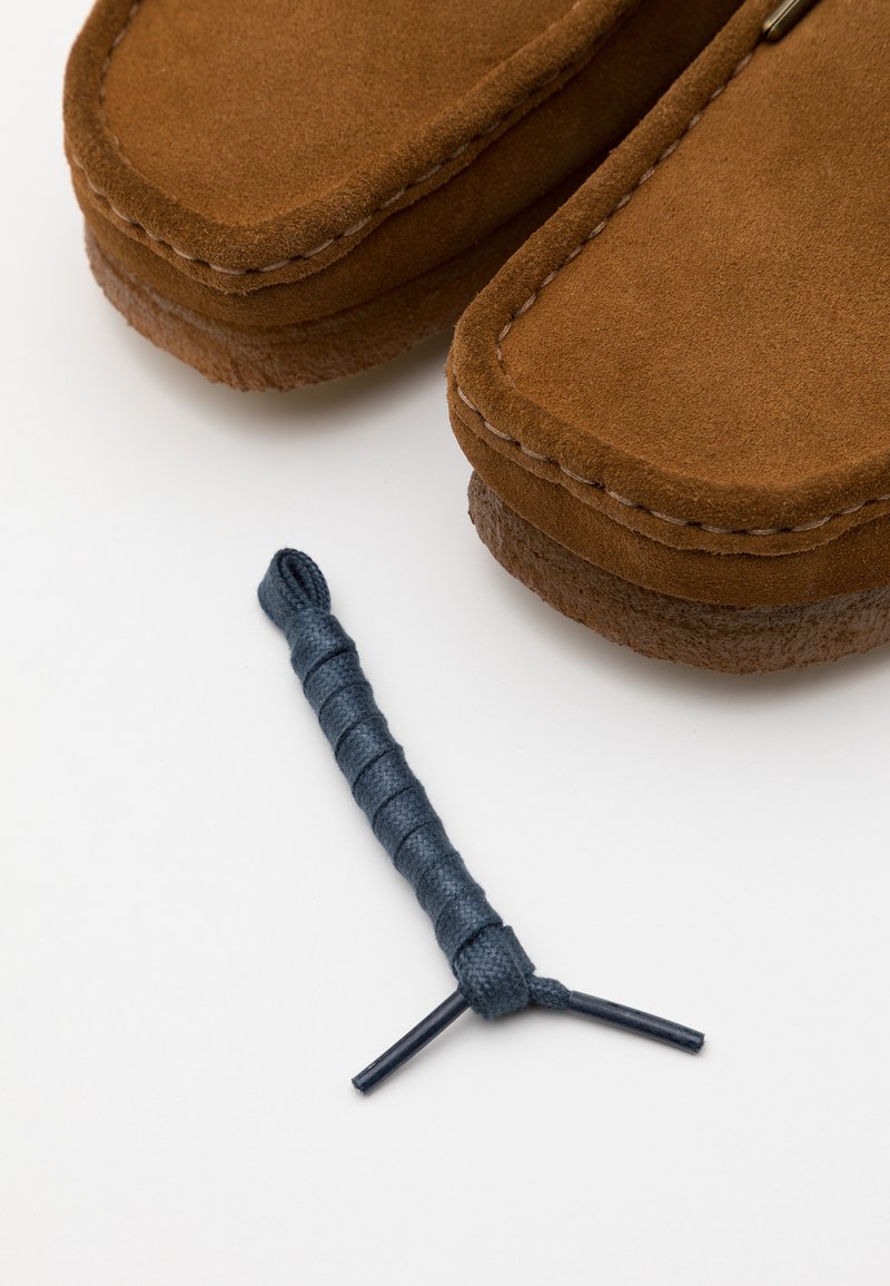 Tan suede moccasins with exposed stitching and a round toe, paired with a dark blue braided lace lying nearby on a white surface.
