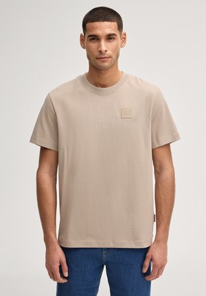 Mann mit kurzen Haaren, der ein beigefarbenes T-Shirt mit Rundhalsausschnitt und blaue Jeans trägt, vor einem schlichten hellen Hintergrund steht.