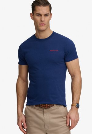 Homme portant un t-shirt bleu marine ajusté avec un texte rouge "Superdry & Co", un pantalon beige, une ceinture tressée marron et une montre-bracelet avec un bracelet marron.