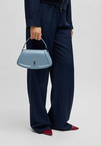 Lichtblauwe leren handtas met een gestructureerde silhoutte, gebogen handvat en zilveren hardwaredetails. G draagt het bij een marineblauwe broek en rode puntige schoenen.