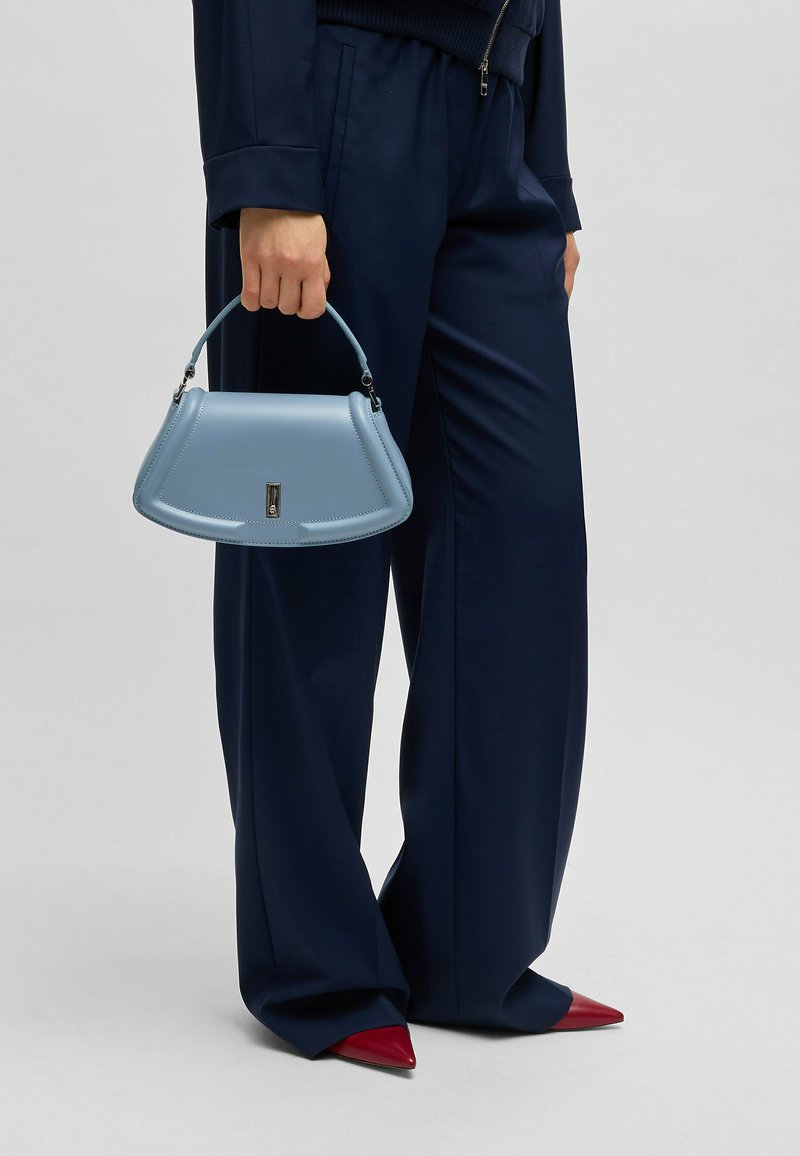 Lichtblauwe leren handtas met een gestructureerde silhoutte, gebogen handvat en zilveren hardwaredetails. G draagt het bij een marineblauwe broek en rode puntige schoenen.