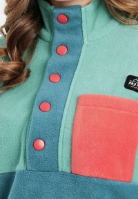 Lekki polarowy pulower w zielono-niebieskim kolorze, z kontrastową koralową kieszenią, różowymi zatrzaskami i wysokim kołnierzem.