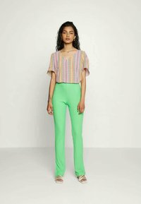 Blusa estampada colorida de mangas curtas combinada com calças justas de malha verde, complementada por sandálias claras.